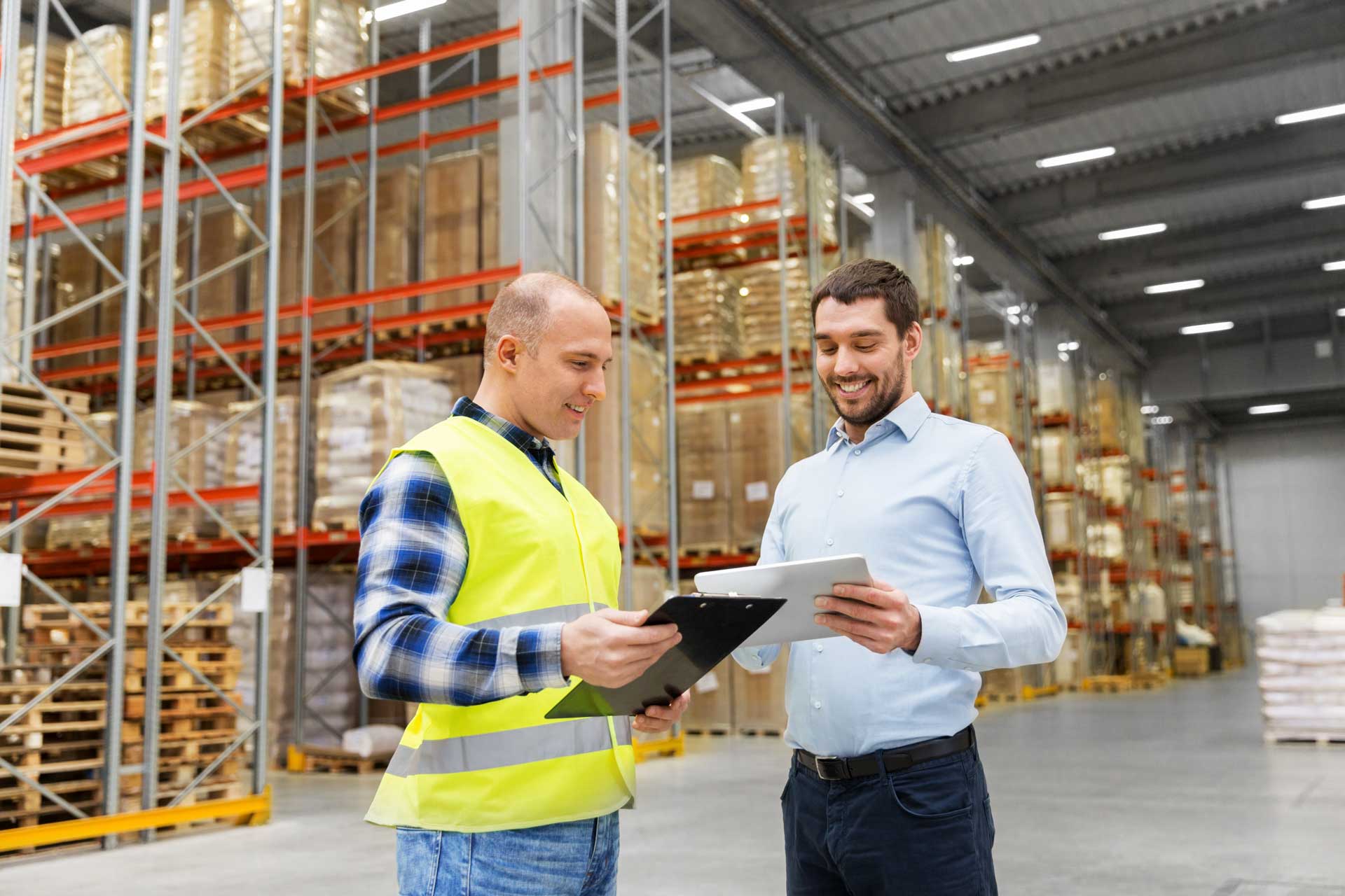 Two men discussing inventory in a warehouse, one wearing a reflective vest holding a clipboard and the other holding a tablet.